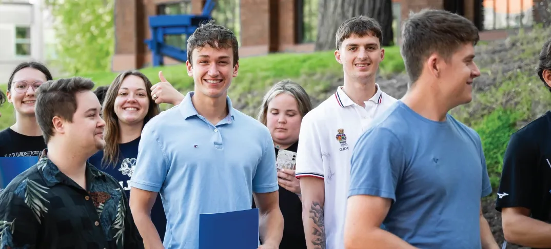 Students smiling at the camera and waiting in a line outdoors on a sunny day.